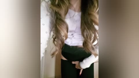 Video of a young woman with long, wavy brown hair, wearing a white cardigan, black pants, and a white top, standing in a doorway with beige walls.