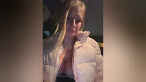 Video of a blonde woman in a white puffy jacket, partially unzipped to reveal a cleavage, with a blurred background.