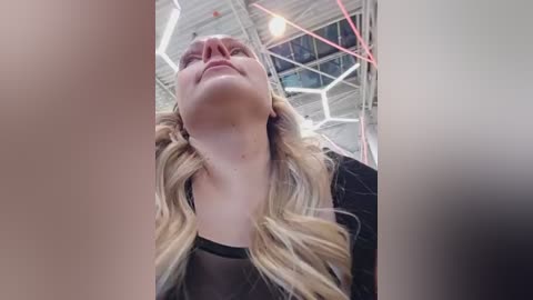 Video of a blonde woman with fair skin and a slight smile, wearing a black top, looking upward in an industrial, high-ceilinged room with exposed pipes and beams.