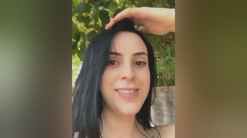 Media: Video of a young woman with long black hair, smiling, with a man's hand on her head, set outdoors with lush green foliage in the background.