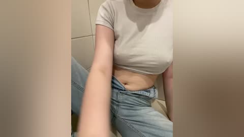 Video of a light-skinned woman with a slender build, wearing a tight white crop top and light-wash jeans, seated in a beige-tiled bathroom. Her midriff is visible, and she is holding the camera, capturing a selfie.