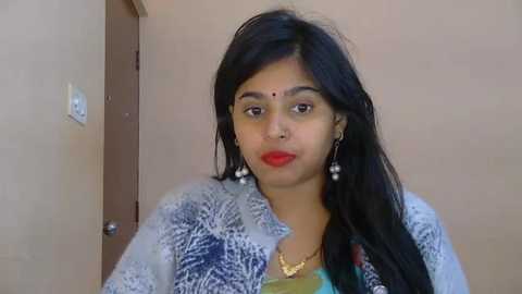 Video of a South Asian woman with long black hair, wearing a blue-patterned blouse, red bindi, and gold earrings. She stands in a simple room with beige walls and a wooden door.