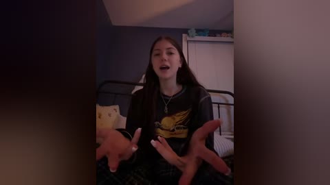 Media: Video of a young woman with long brown hair, wearing a black graphic tee, sitting on a bed in a dimly lit room.