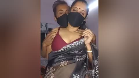 A video of two women wearing black masks, one in a maroon bra, the other in a black saree with intricate patterns, embracing closely in a dimly lit room.