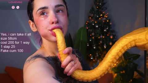 A photo of a young woman with dark hair, wearing a yellow, snake-like costume, biting a banana, in a dimly-lit room with a decorated Christmas tree.