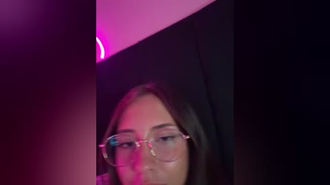 A video of a young woman with fair skin and glasses, wearing a black hijab, partially visible under a neon pink light.