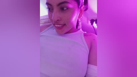 Video of a young woman with light brown skin, dark hair, and brown eyes, wearing a white ribbed tank top, smiling under purple lighting, her arm bandaged.