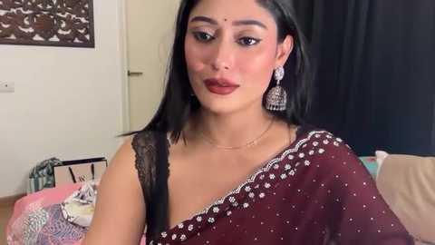 Video of a South Asian woman with medium skin tone, long black hair, and maroon saree adorned with white sequins. She wears a bindi, red lipstick, and dangling earrings. Background includes a beige wall, black curtain, and colorful fabric on the left.