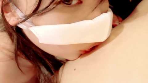 Close-up video of a young Asian woman with brown hair, wearing a white bandage over her mouth, looking up with a serious expression, partially visible breasts, and a blurred background.