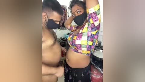 Video of a shirtless man with a tattooed arm and a woman in a colorful crop top, black mask, and pants, in a cluttered room.