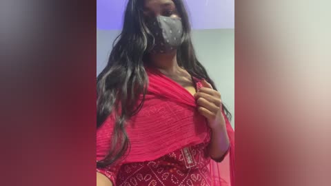 A video shows a woman with long black hair, wearing a red sari with a white pattern, a face mask, and a headband, indoors against a purple and white backdrop.