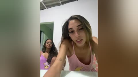 A video of two young women in a bathroom, one with long brown hair and a pink tank top, another with long black hair and a purple top.
