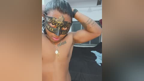Video of a shirtless, muscular, dark-skinned man with long hair, wearing a black Venetian mask and a necklace, posing in a bedroom with a bed and windows.