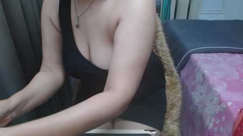 Video of a light-skinned woman with medium-sized breasts, wearing a black, low-cut top, sitting on a car seat. Her arms are resting on a dashboard, with a pink floral-patterned blanket and a beige fur-trimmed garment in the background.