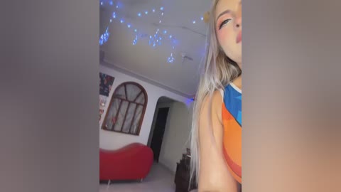 Media: Video of a young woman with long, platinum blonde hair, wearing a bright orange and blue sleeveless top, in a dimly lit, cozy living room with red sofa, blue string lights, and framed artwork on the wall.