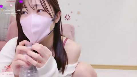 A video of an Asian woman with long brown hair, wearing a pink face mask, white long-sleeve top, and a red bra, sitting indoors.