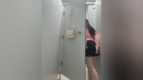 Video of a woman in a public restroom stall, wearing a pink top and black shorts, carrying a beige handbag. The stall has a grey door and beige floor, with a mirror reflecting her back.
