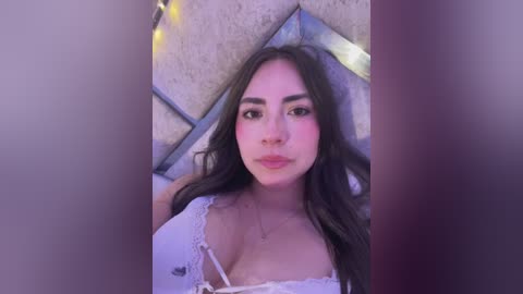 Video of a young woman with long, wavy brown hair, fair skin, and a slender physique. She wears a white lace top, revealing cleavage. Background features a geometric, light-colored wall and fairy lights.