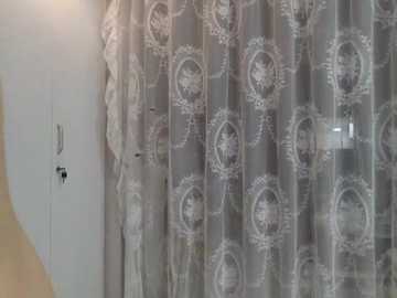 Media: Video of a bedroom featuring a sheer, patterned lace curtain with an ornate floral design. The curtain is partially open, revealing a white door with a metal handle on the left. The room has a soft, cozy ambiance with a light wooden floor.
