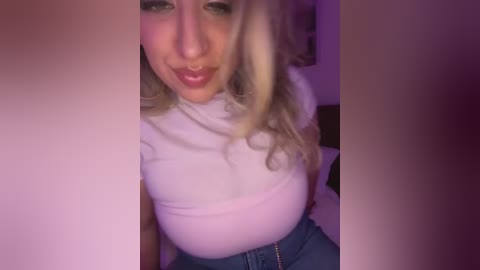 Video of a young woman with blonde hair, fair skin, and large breasts, wearing a white turtleneck and blue jeans, smiling. Background is dimly lit with purple hues.