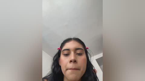 Video of a young woman with long black hair, wearing red hair clips, gazing downward. She has medium brown skin and a serene expression. The background is a plain white ceiling.