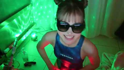 Video of a smiling young Asian woman in a blue swimsuit, wearing black sunglasses and bunny ears, in a dimly lit room with green neon lights, cluttered with random items on the floor.