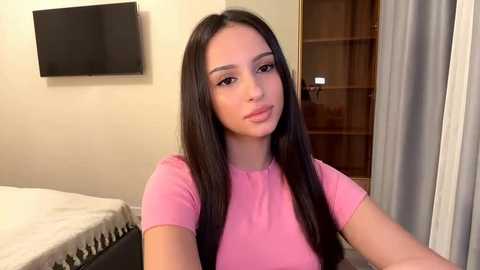 Video of a young woman with long, straight, dark hair, wearing a pink t-shirt, sitting indoors in a bedroom with a flat-screen TV, beige walls, and a wooden closet in the background.