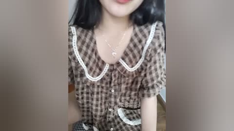 Video of a young woman with pale skin, straight black hair, and a petite frame. She wears a brown plaid dress with a white lace collar and a silver necklace. The background is a plain beige wall.