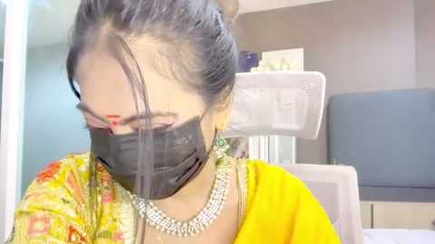 Video of an Indian woman in a yellow saree with a black mask, seated in an office, wearing a silver necklace. Background shows a white chair, a gray wall, and a blue table.