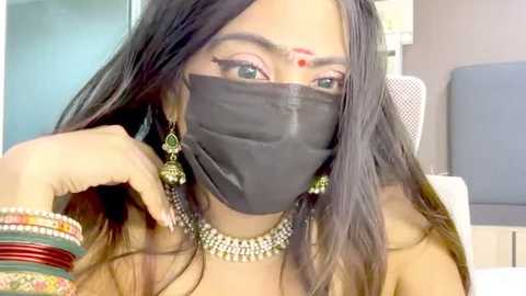 Video of a South Asian woman with long black hair, wearing a black mask, red bindi, gold jewelry, and colorful bangles, seated indoors.