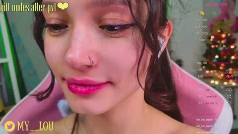 Media: Video of a young woman with fair skin, brown hair, and a nose ring, wearing pink lipstick, taken in a room with a Christmas tree in the background.