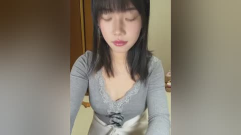 Video of a young Asian woman with straight black hair, wearing a grey long-sleeved top with lace trim, standing indoors.