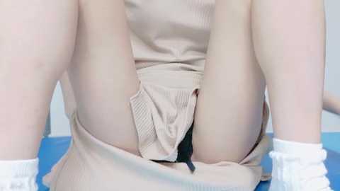 Video of a person's legs in a beige skirt, partially exposing black underwear, sitting on a blue surface with white socks.