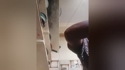 Video of a person with a muscular build, wearing a dark shirt, standing in a cluttered room with shelves holding various objects, including colorful bowls. The background shows a beige wall and a framed picture.