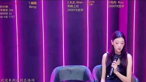 Video of a female singer with long black hair, dressed in a black sleeveless top, holding a microphone, against a vibrant purple backdrop with LED lights and numbered panels.