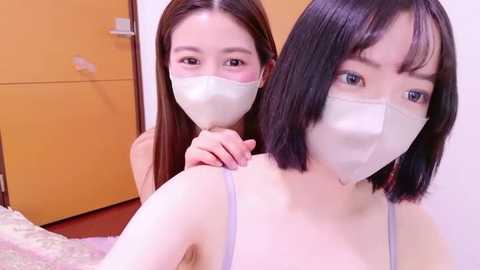Media: Video of two young East Asian women with light skin, wearing face masks, in a cozy bedroom with wooden doors and a plush carpet.