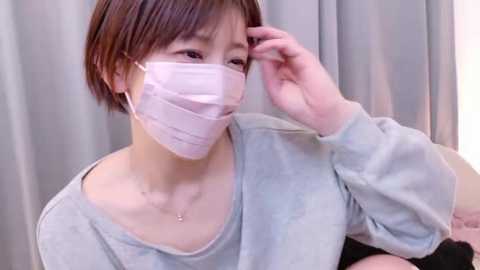 Video of a young Asian woman with short brown hair, wearing a light gray sweater and a white surgical mask, adjusting her glasses with her right hand, against a light gray curtain background.
