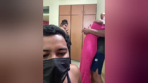 Video of a man wearing a black face mask, taking a selfie with a woman in a red dress. Background shows wooden cabinets and a woman in a pink dress.