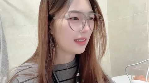 A close-up video of an East Asian woman with long, straight brown hair and light skin, wearing large, round glasses and a gray shirt. She is smiling and appears to be in a bathroom with tiled walls.