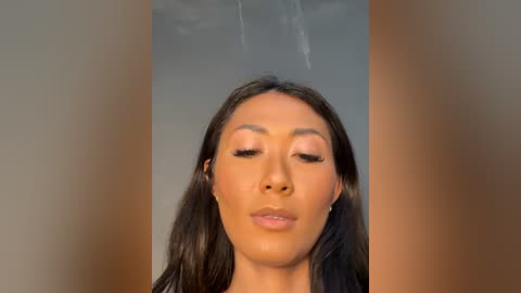 Video of a young Asian woman with long black hair, wearing makeup with pink eyeshadow, closed eyes, and a neutral expression. The background is blurred with soft lighting, emphasizing her face.