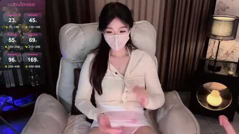 A video of an East Asian woman with long black hair, wearing a white face mask and long-sleeve top, sitting in a beige armchair, holding a controller, in a dimly lit room with a digital interface showing statistics.