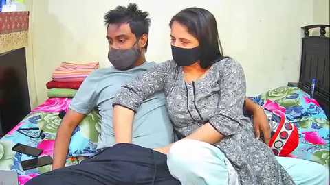 Media: Video of a man and woman, both masked, sitting on a colorful bedspread in a modest bedroom. The woman, with long black hair, wears a patterned dress; the man, with spiky black hair, wears a light gray t-shirt.