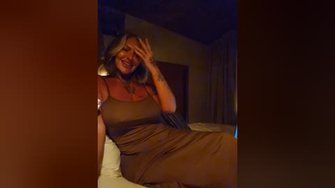 Media: Video of a smiling, middle-aged woman with long, wavy blonde hair, wearing a form-fitting, sleeveless brown dress, seated on a bed in a dimly lit, cozy bedroom with wooden ceiling beams.