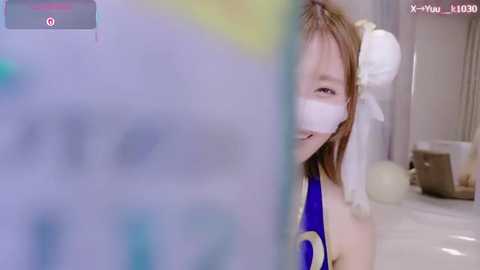 A video of a young woman with light skin, wearing a blue and white Chun-Li cosplay outfit with a white mask covering her eyes and nose, smiling while holding a phone in front of a mirror.