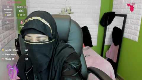 Video of a woman wearing a black hijab and dark glasses, sitting in a chair, with a green wall and white brick backdrop, and a pink blanket on her lap.