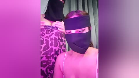 Video of a woman with light skin, wearing a black mask, leopard print dress, and a thin strap top, standing in front of a mirror, with purple and pink lighting.