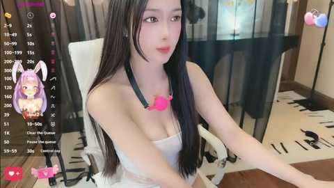 A video of an East Asian woman with long black hair, wearing a strapless white top, sitting at a desk. The background features a modern room with a white chair and black-and-white patterned rug. A pink bunny filter overlays her face.