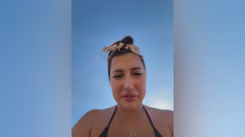 Video of a woman with medium-brown skin, dark hair tied up with a beige headband, wearing a black bikini top, gazing at the camera against a clear blue sky.