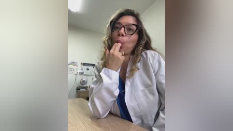 Video of a Caucasian woman with wavy blonde hair, wearing glasses and a white lab coat, biting her finger pensively at a desk in a clinical setting.