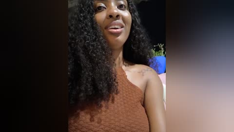 Video of a smiling Black woman with a medium brown complexion and large, voluminous curly black hair. She wears a one-shoulder, brown lace top. The background is a dark gradient with a blue potted plant.